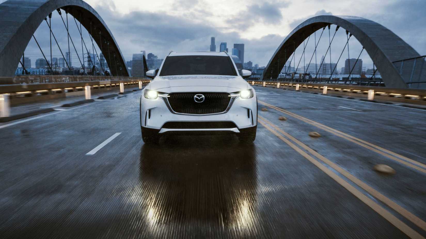 Dynamic front-end shot of a white 2025 Mazda CX-90 driving over a bridge with a cityscape in the background.