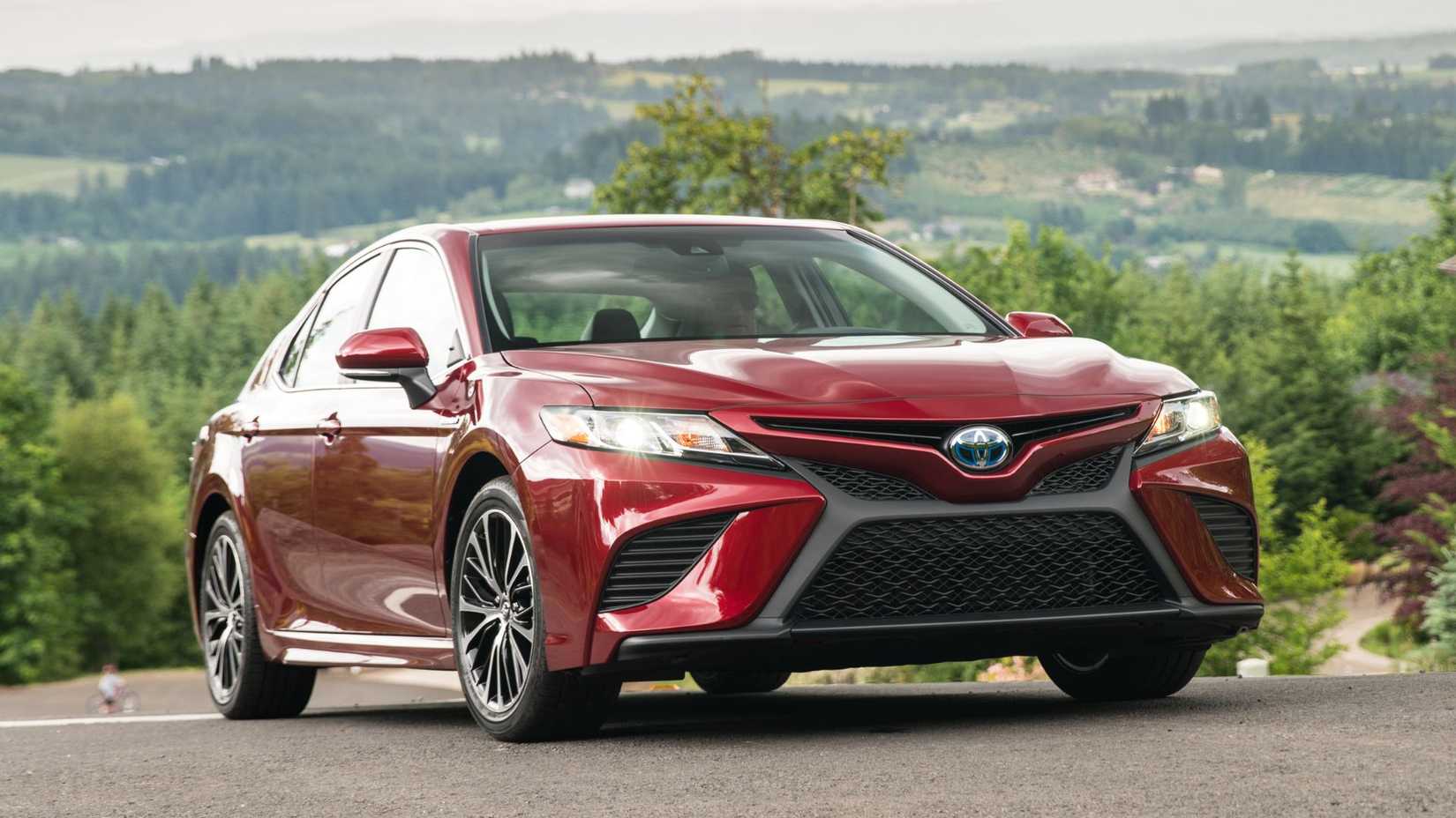 পটভূমিতে ক্ষেত্র সহ একটি লাল 2020 Toyota Camry SE হাইব্রিডের স্ট্যাটিক সামনের 3/4 শট।
