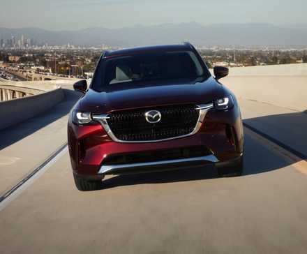 Dynamic front-end shot of a red 2025 Mazda CX-90 driving on a freeway with a cityscape in the background.