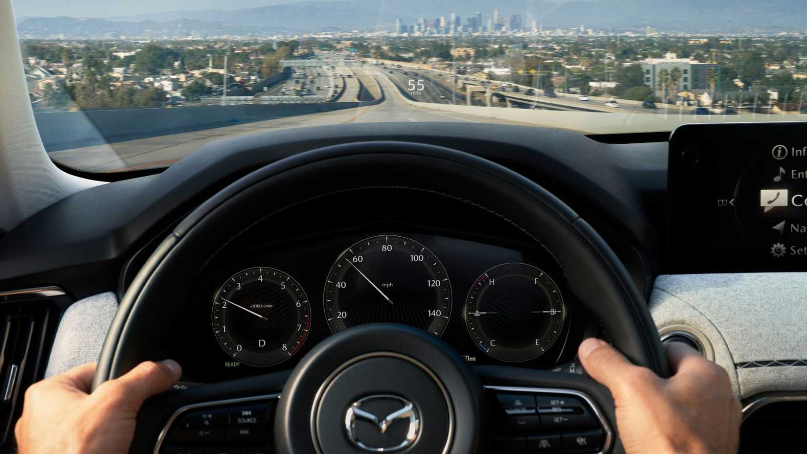 Close-up shot of a man's hands on the steering wheel of a 2025 Mazda CX-90, with a citscape in the background.