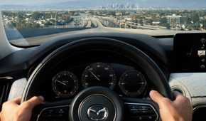 Close-up shot of a man's hands on the steering wheel of a 2025 Mazda CX-90, with a citscape in the background.