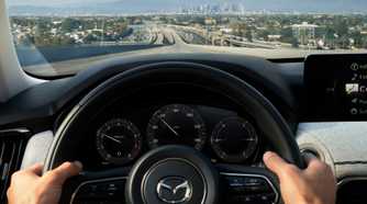 Close-up shot of a man's hands on the steering wheel of a 2025 Mazda CX-90, with a citscape in the background.