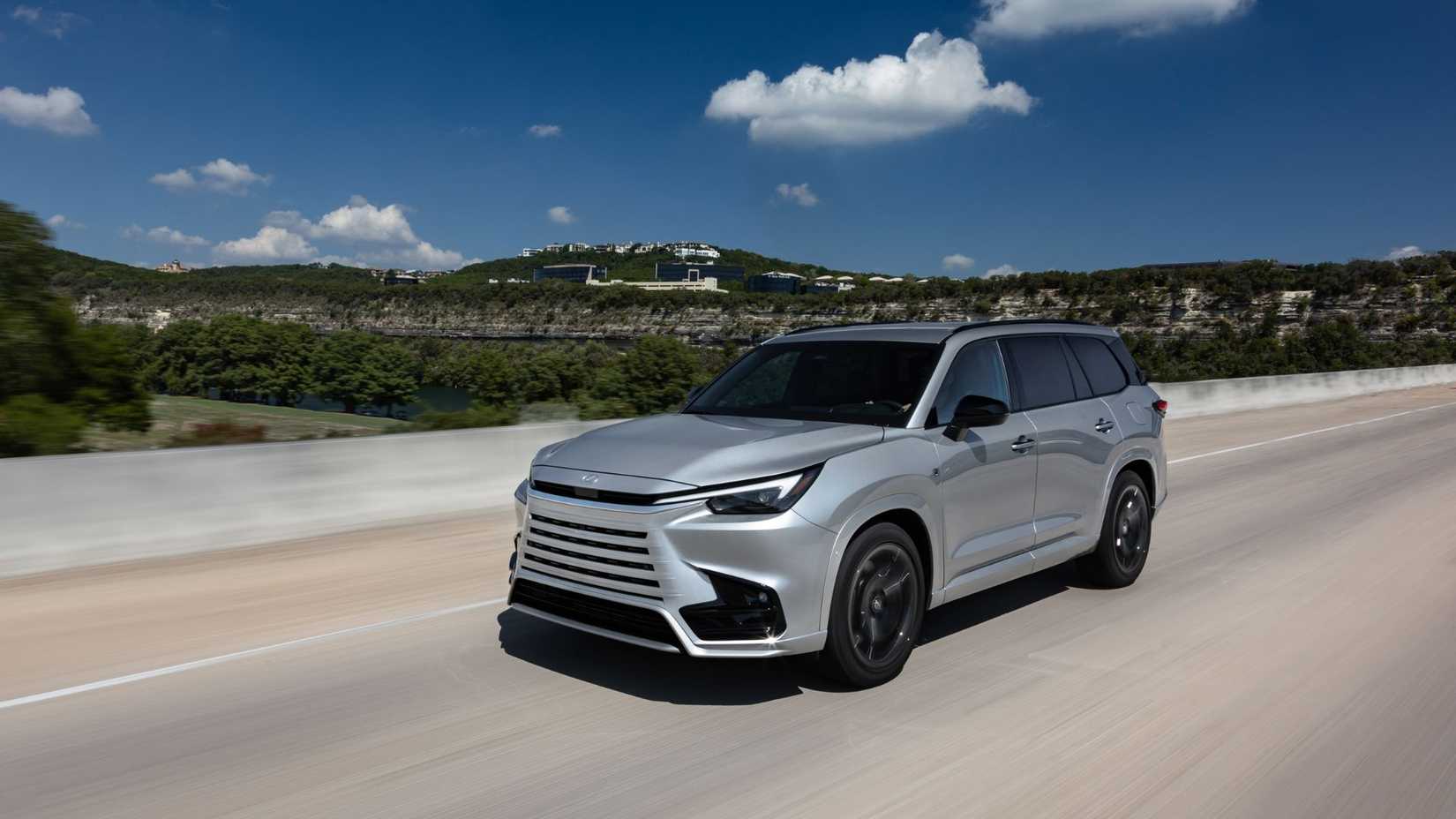 Dynamic front 3/4 shot of a silver 2025 Lexus TX 500h FSPORT Premium driving on a country road with hills in the background.