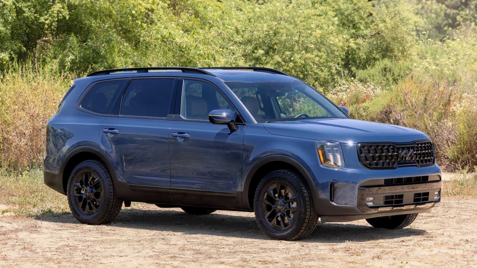 Foto estática frontal 3/4 de um Kia Telluride 2025 azul estacionado na areia com árvores ao fundo.