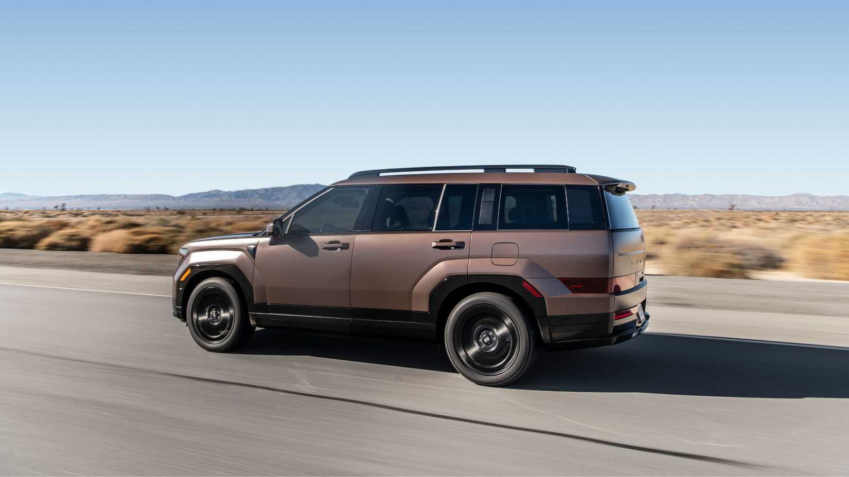 Dynamic side profile photo of a bronze 2025 Hyundai Santa Fe Hybrid driving on a country road through the desert.