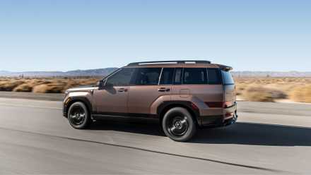 Dynamic side profile shot of a bronze 2025 Hyundai Santa Fe Hybrid driving on a country road through the desert.