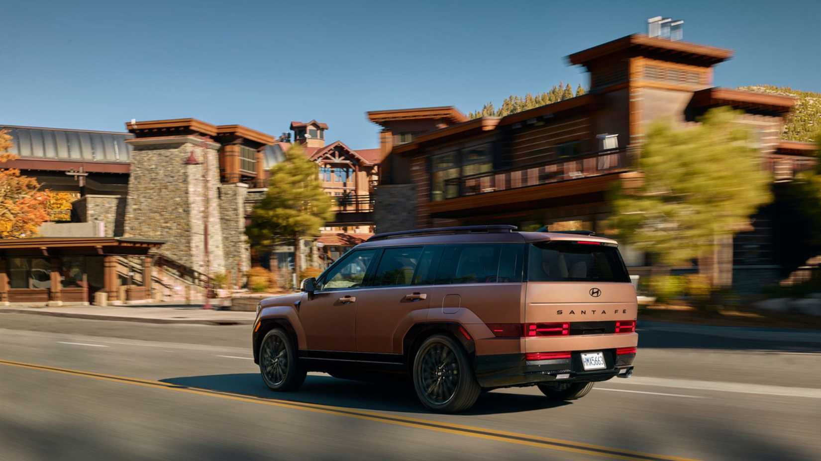 Dynamic rear 3/4 shot of bronze 2025 Hyundai Santa Fe Hybrid driving around town.