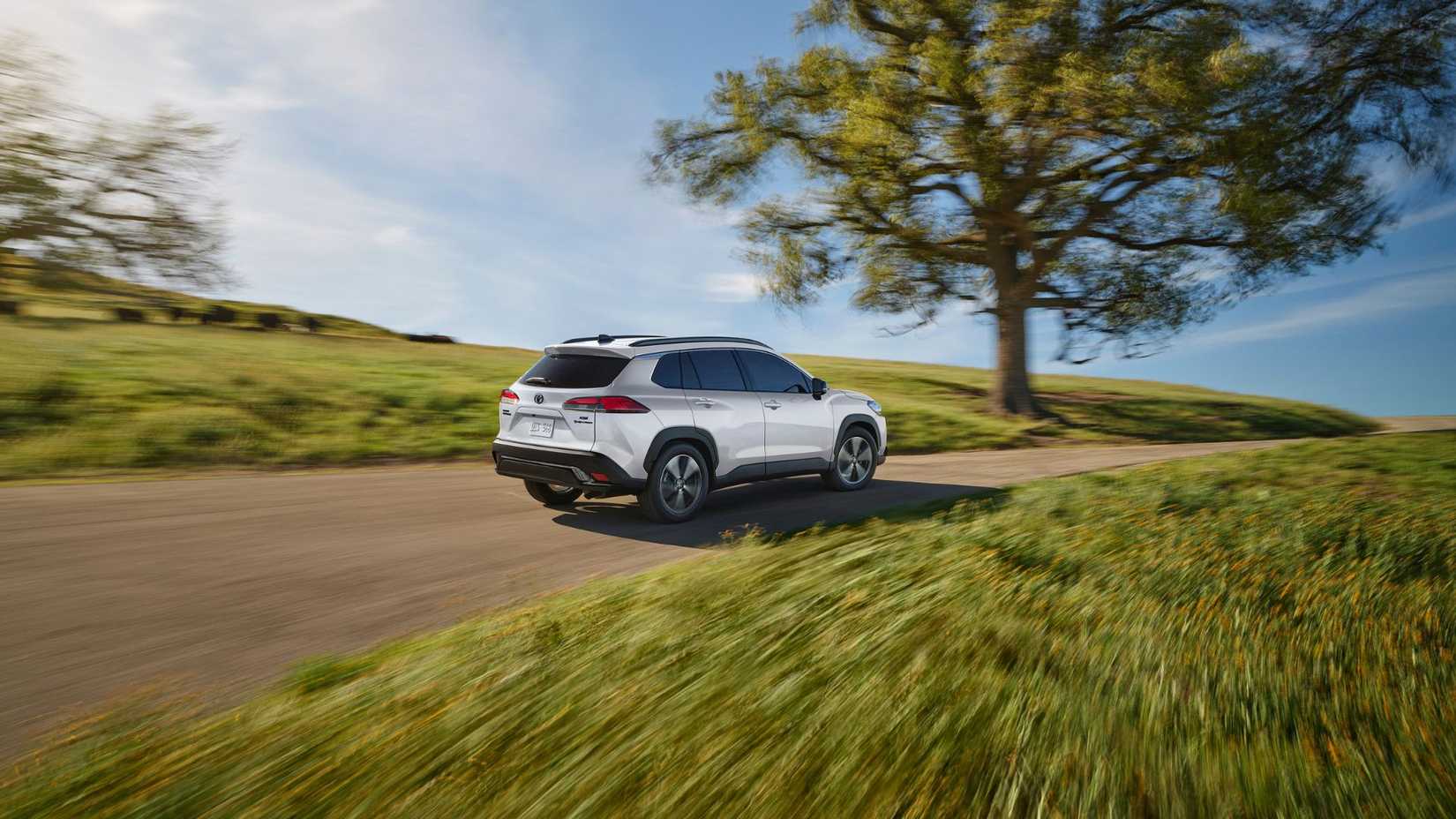 Foto traseira dinâmica 3/4 de um Toyota Corolla Cross Hybrid XSE 2025 branco dirigindo em uma estrada estreita com uma grande árvore ao fundo.