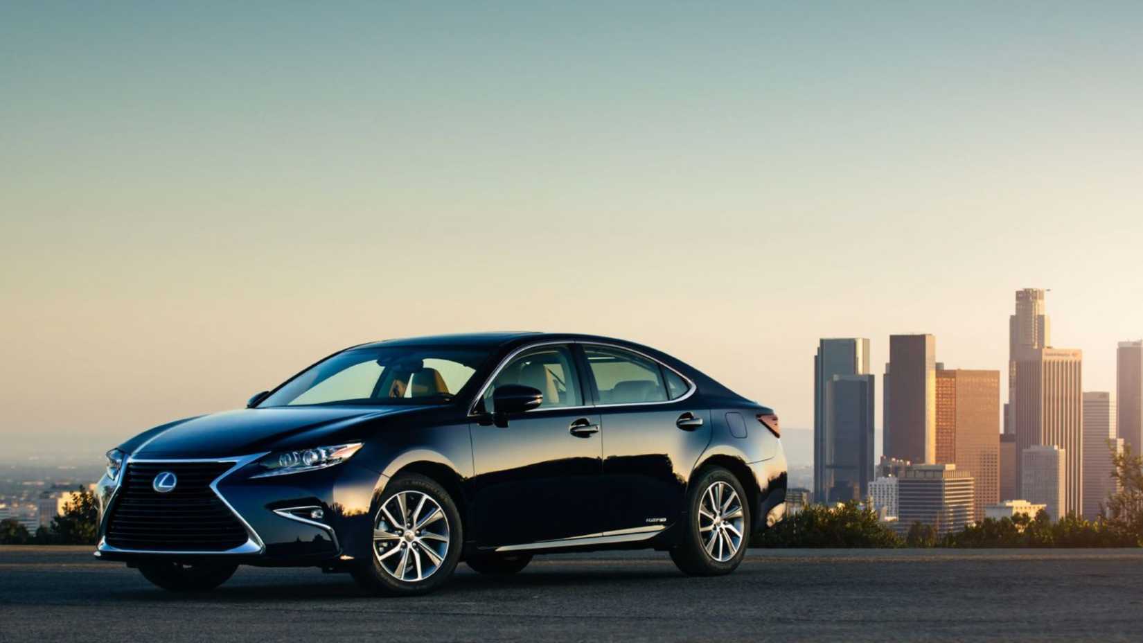 Static front 3/4 shot of a black 2016 Lexus ES 300h parked on tarmac with a cityscape in the background.
