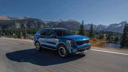Dynamic front 3/4 shot of a blue 2025 Kia Sorento Hybrid driving on a country road with snowcapped mountains in the background.
