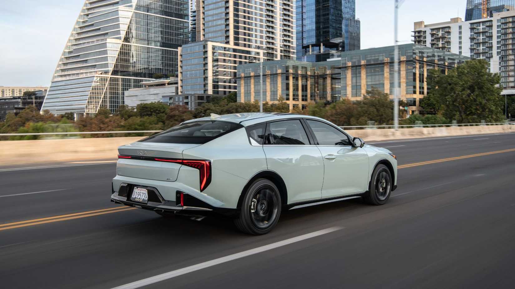 Plan 3/4 arrière dynamique d'une Kia K4 2025 grise conduisant en ville.