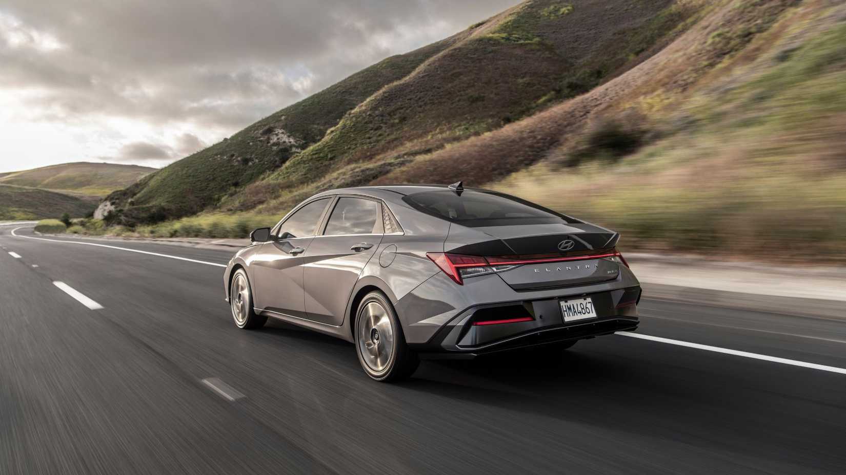 Dynamic rear 3/4 shot of a gray 2025 Hyundai Elantra drivinhg on a country road with hills in the background.