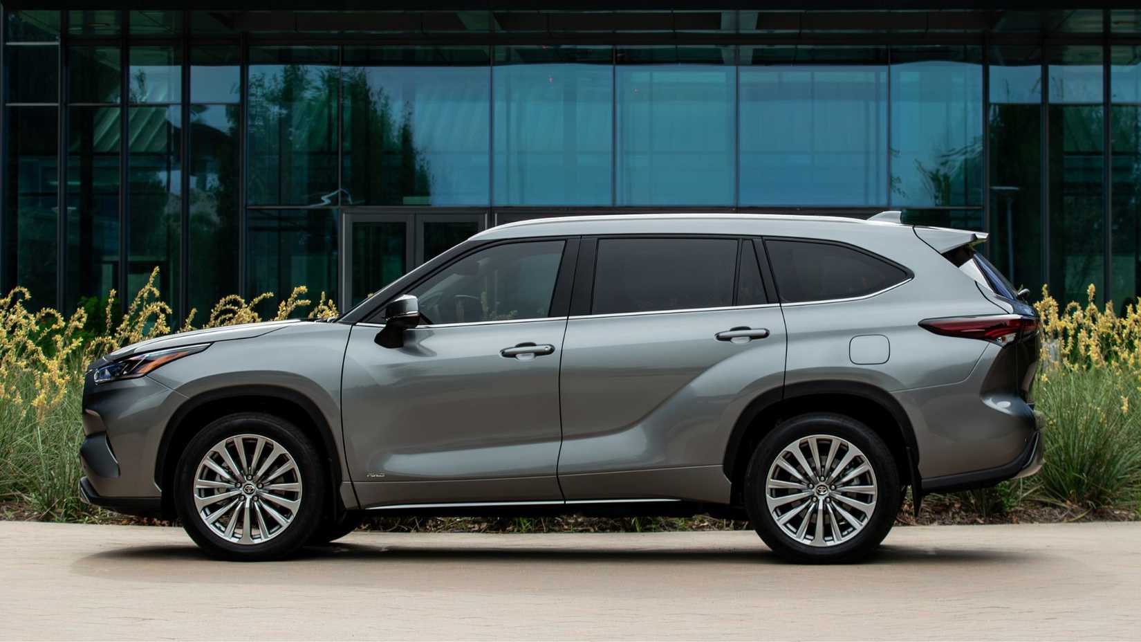 Static side profile shot of a gray 2025 Toyota Highlander Hybrid parked outside of a modern glass-fronted building.