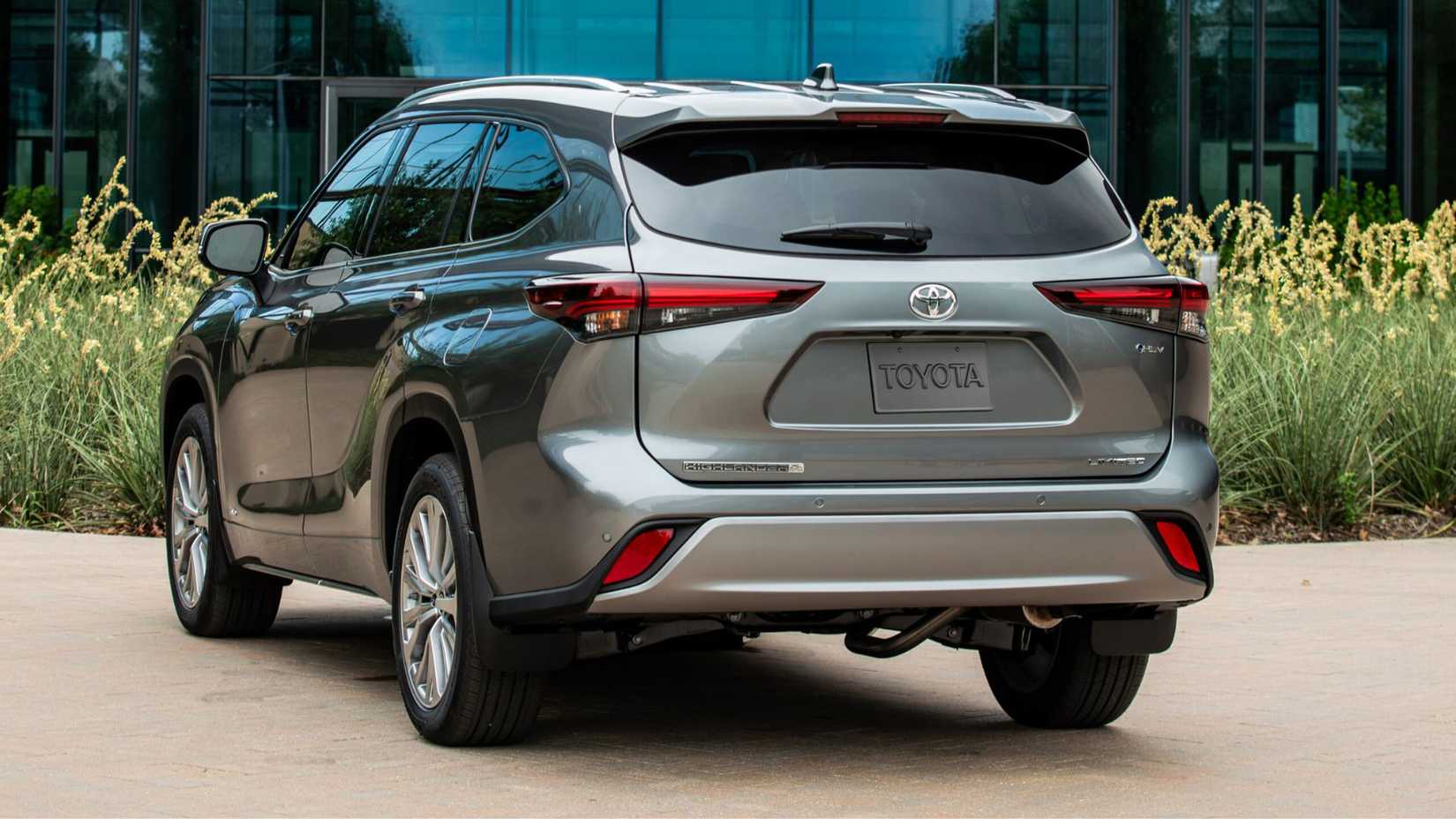 Static rear 3/4 shot of a gray 2025 Toyota Highlander Hybrid parked in front of a modern glass-fronted building.