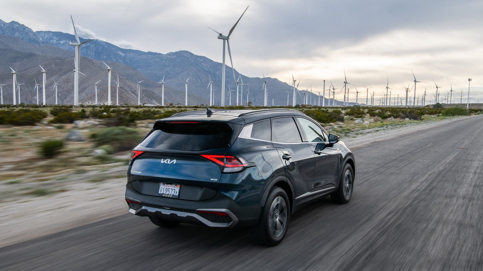 Foto traseira dinâmica 3/4 de um Kia Sportage Hybrid 2025 azul dirigindo em uma estrada secundária com montanhas e turbinas eólicas ao fundo.