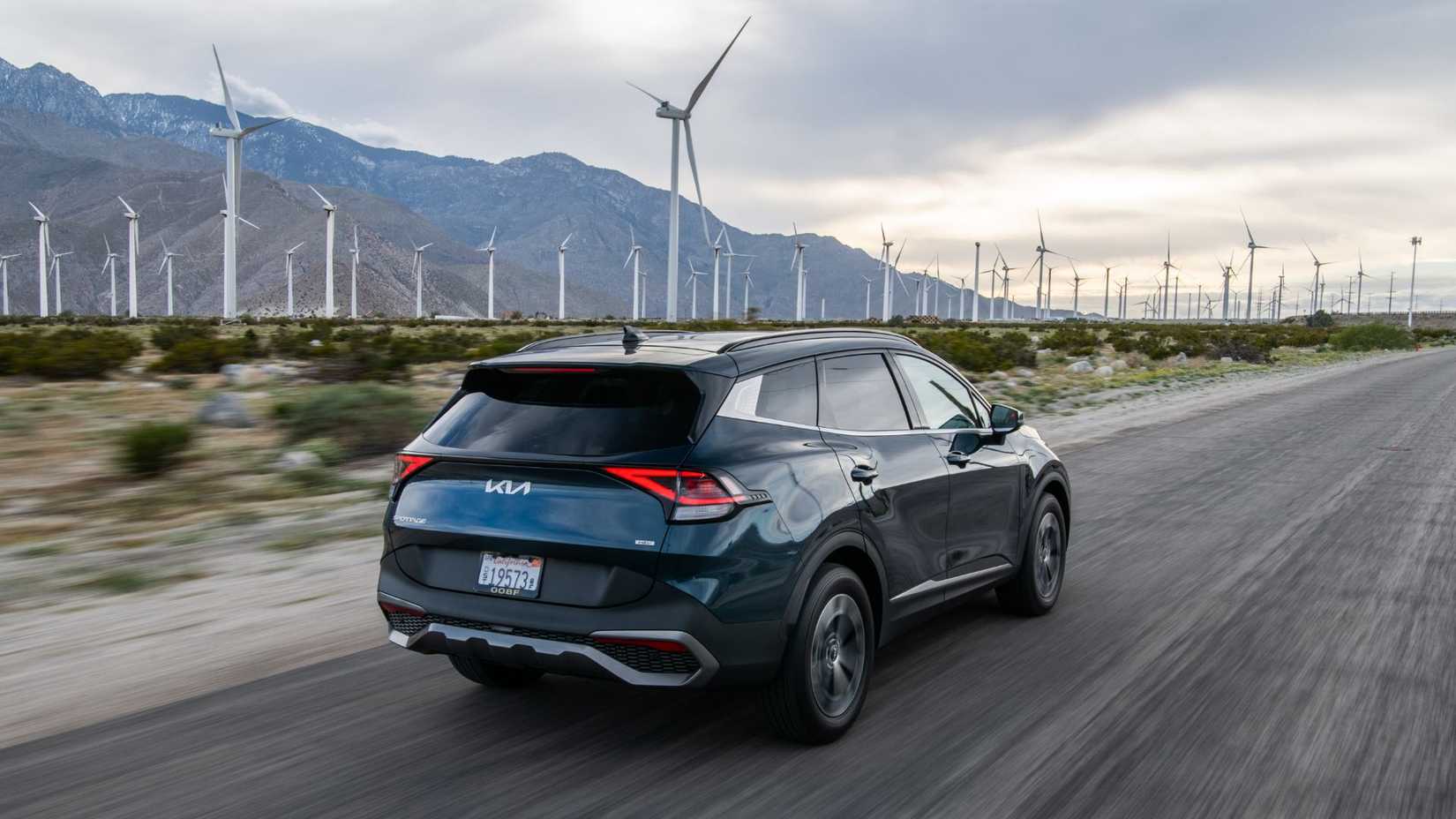 Toma dinámica de 3/4 de la parte trasera de un Kia Sportage Hybrid azul 2025 conduciendo por una carretera rural con montañas y turbinas eólicas de fondo.