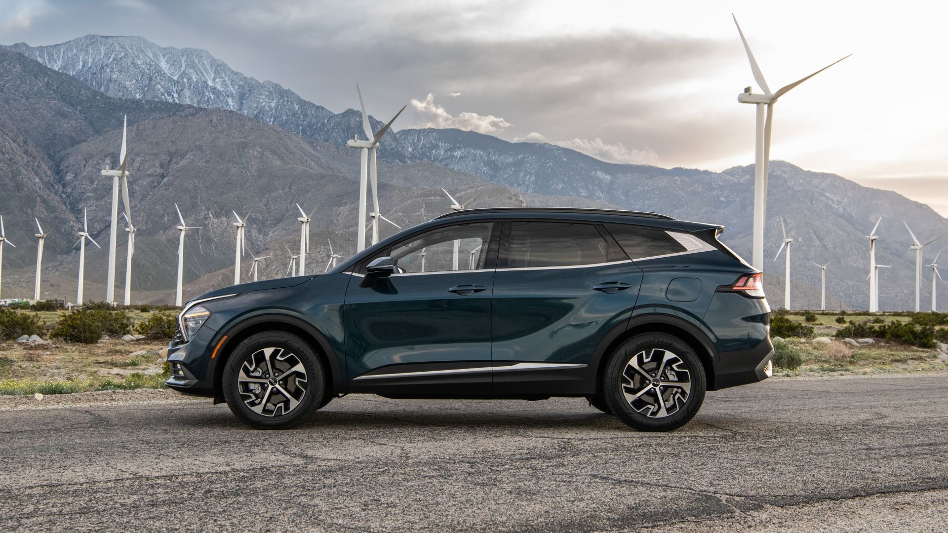 Статический боковой снимок серого Kia Sportage Hybrid 2025 года выпуска с горами и ветряными турбинами на заднем плане.