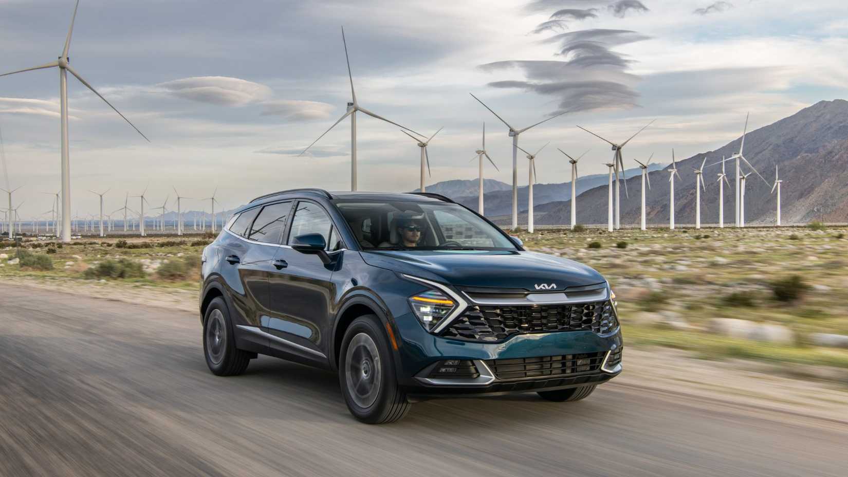 Dynamic front 3/4 shot of a blue 2025 Kia Sportage Hybrid driving on a country road with mountains and wind turbines in the background.