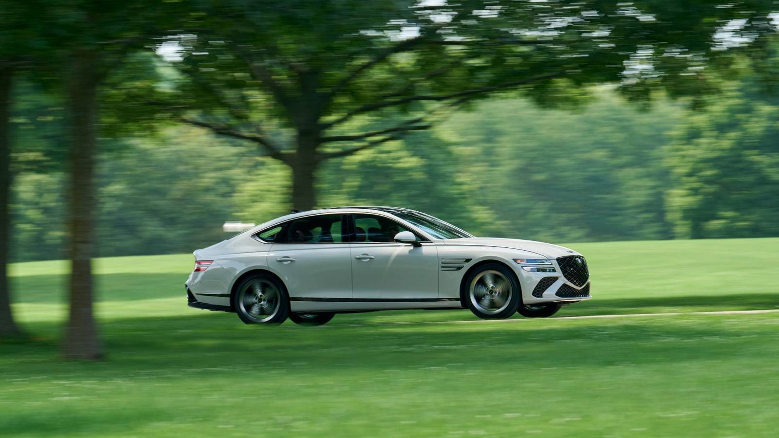Dynamic side profile shot of a gray 2025 Genesis G80 driving on a country road with trees in the background.
