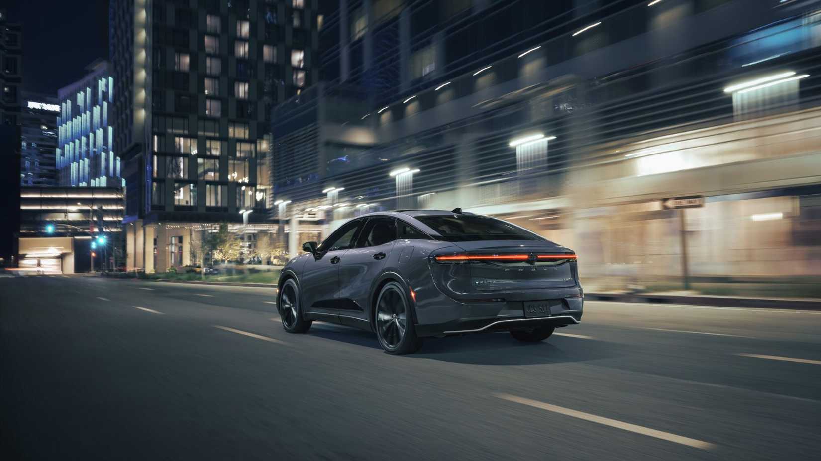 Dynamic rear 3/4 shot of a gray 2026 Toyota Crown Nightshade driving at night in a city.