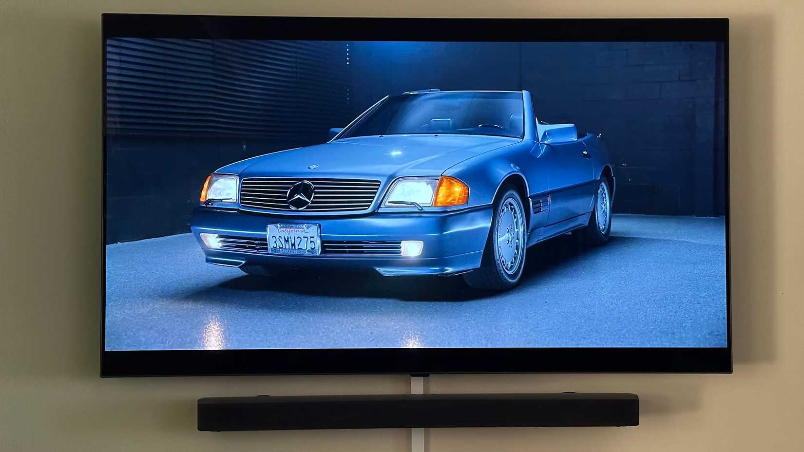 A OLED TV mounted on the wall with a soundbar underneath. The TV shows a scene from the Hagerty YouTube channel featuring a classic Mercedes SL.