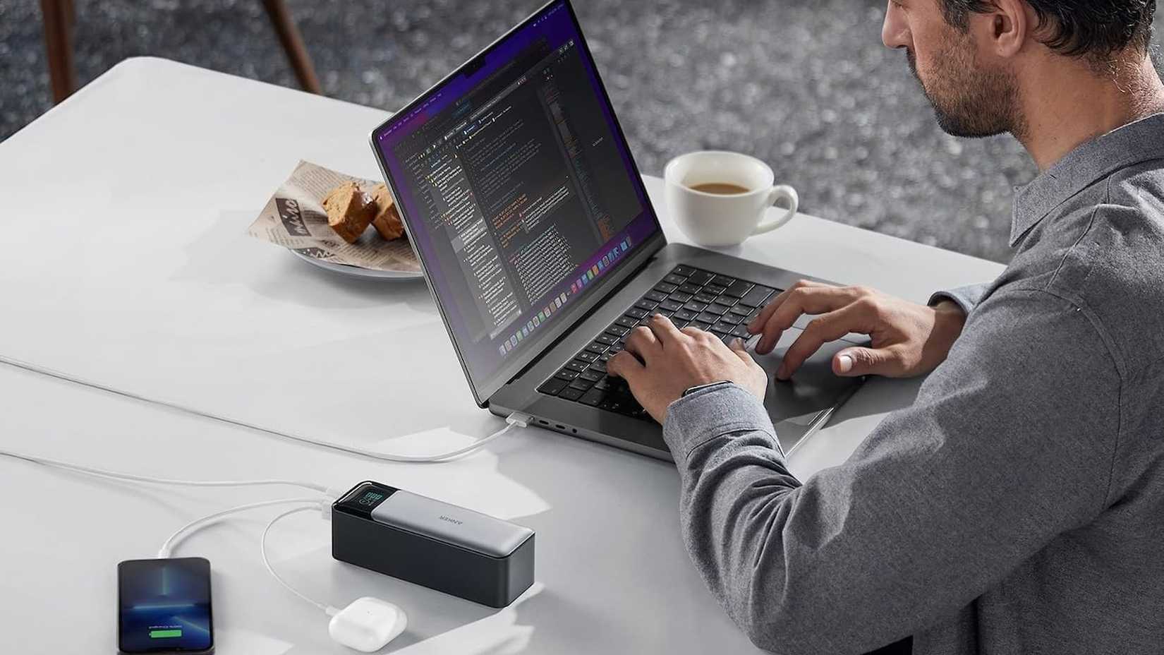 An Anker 737 power bank being used to charge a laptop and phone and earphones while a man works and eats