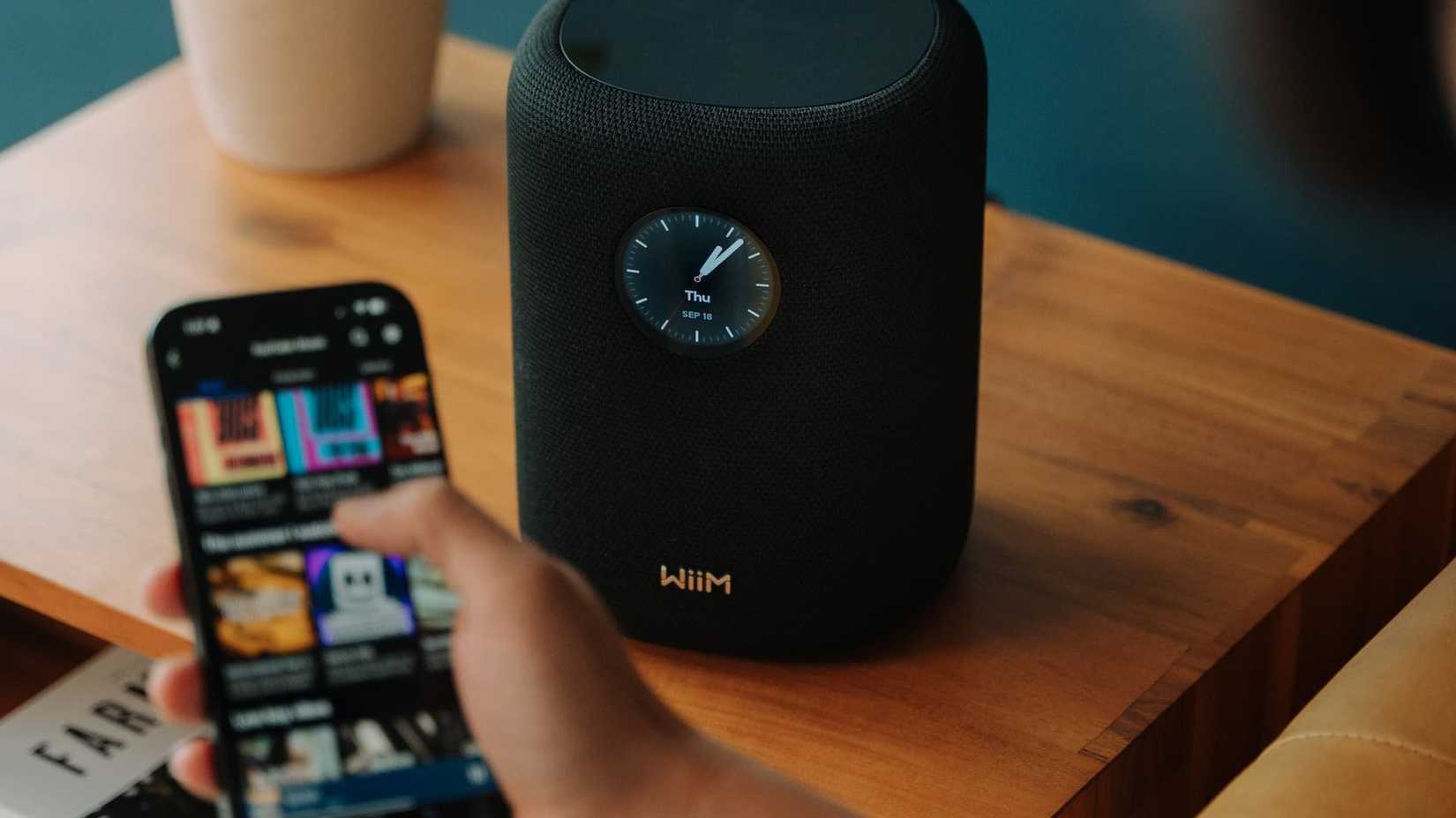 Alguém segurando um telefone e escolhendo uma música ao lado de um WiiM Sound preto mostrando as horas