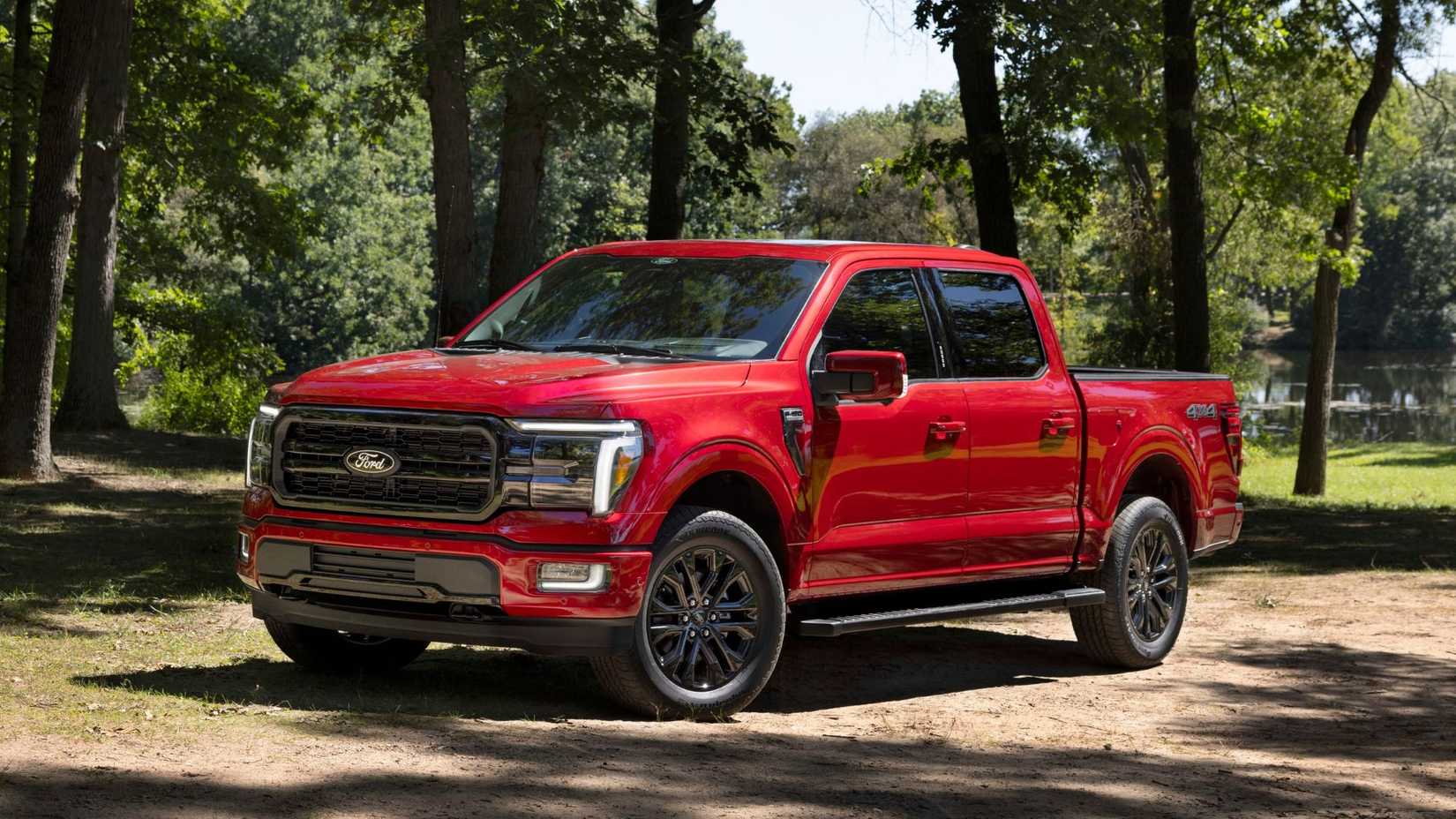 Static front 3/4 shot of a red 2024 Ford F-150 Lariat parked on dirt with trees in the background.