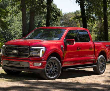 Static front 3/4 shot of a red 2024 Ford F-150 Lariat parked on dirt with trees in the background.