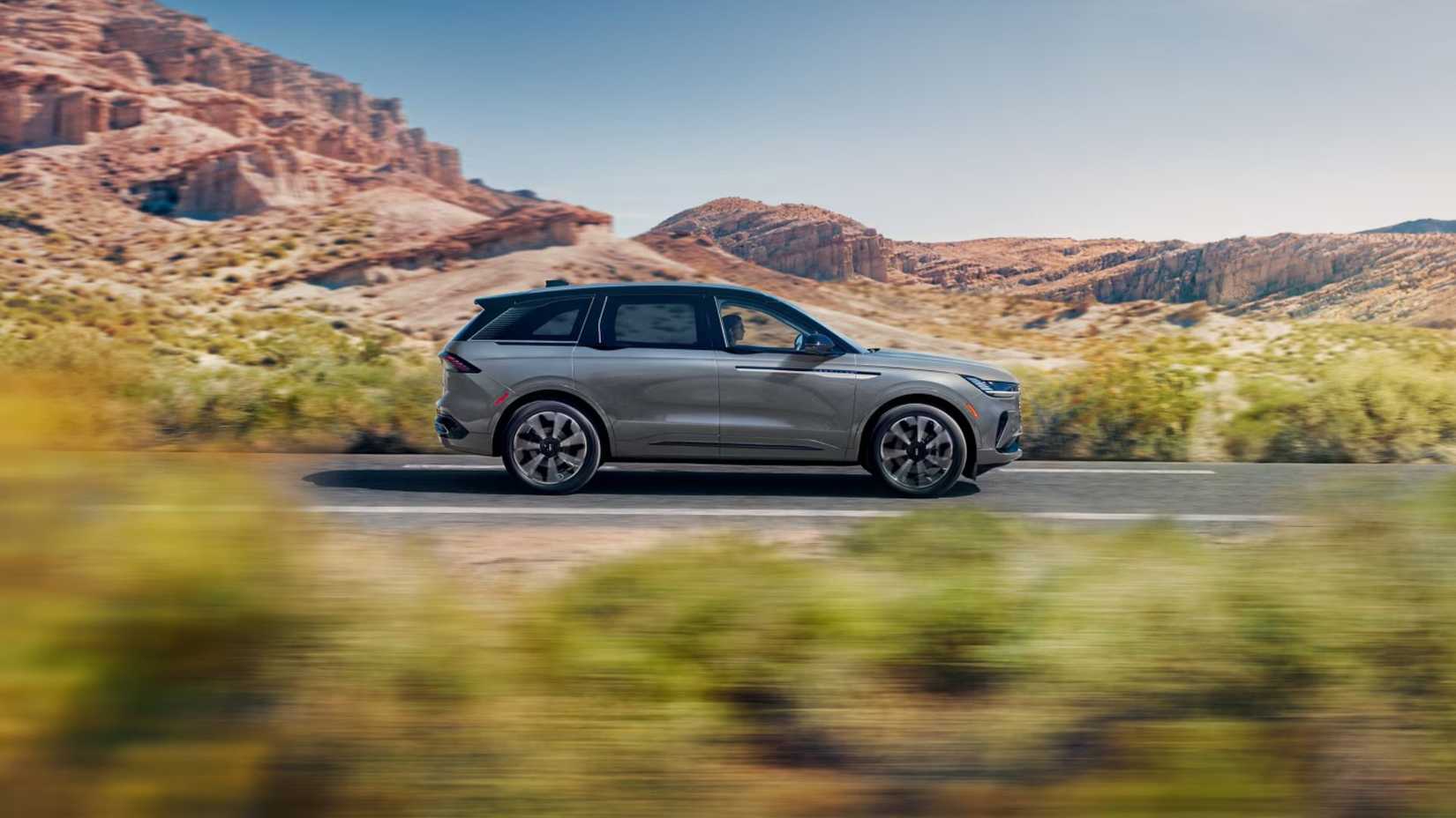 Dynamic side profile shot of a gray 2026 Lincoln Nautilus Hybrid driving on a country road.