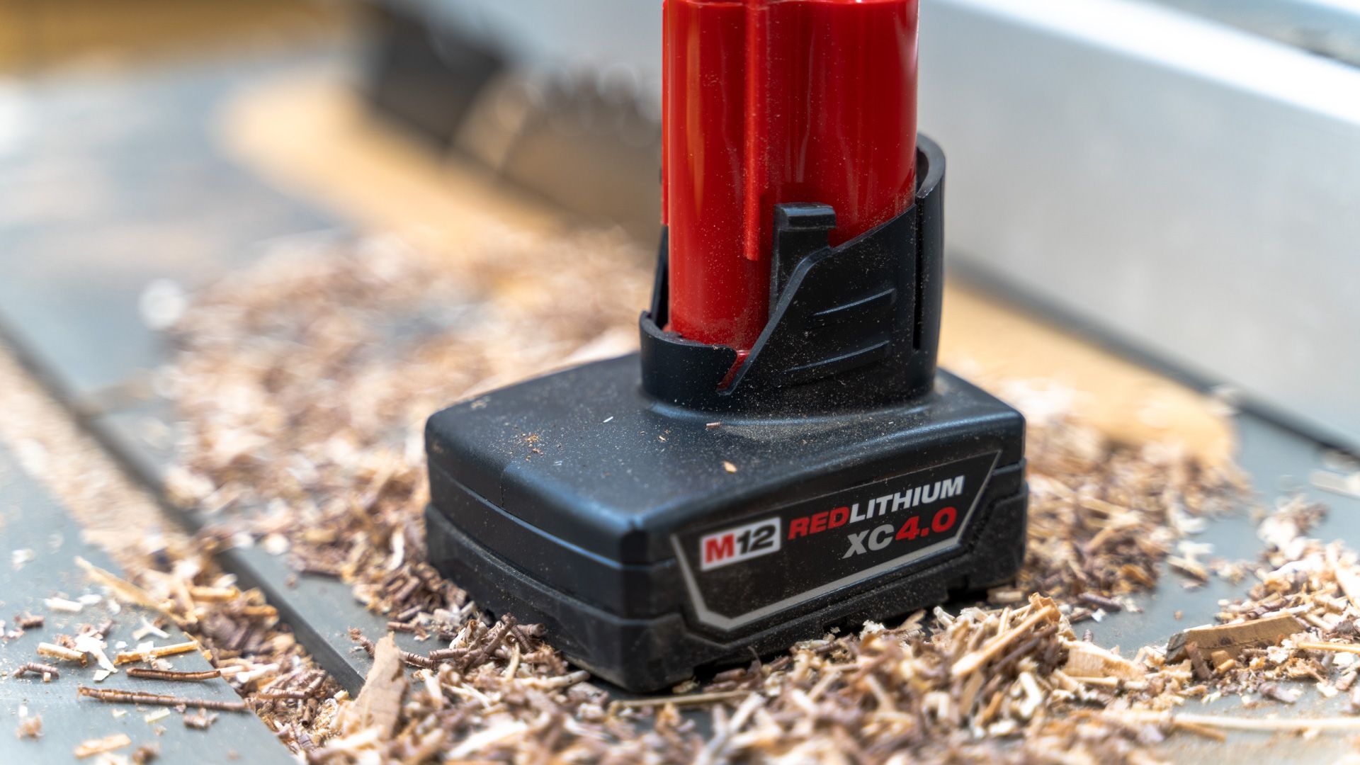 A close up of a Milwaukee M12 battery sitting on a table saw with sawdust around it.