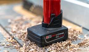 A close up of a Milwaukee M12 battery sitting on a table saw with sawdust around it.