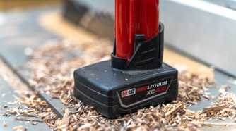 A close up of a Milwaukee M12 battery sitting on a table saw with sawdust around it.