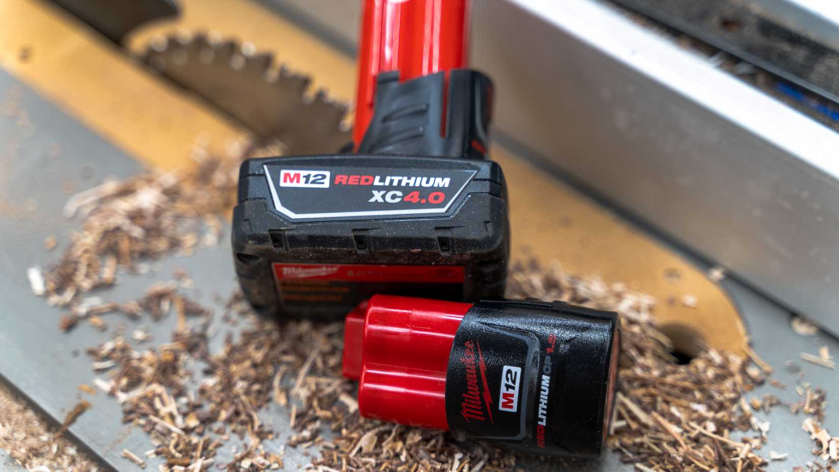 Two Milwaukee M12 batteries sitting on a table saw with sawdust and a blade in the background.
