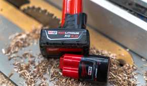 Two Milwaukee M12 batteries sitting on a table saw with sawdust and a blade in the background.