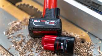 Two Milwaukee M12 batteries sitting on a table saw with sawdust and a blade in the background.