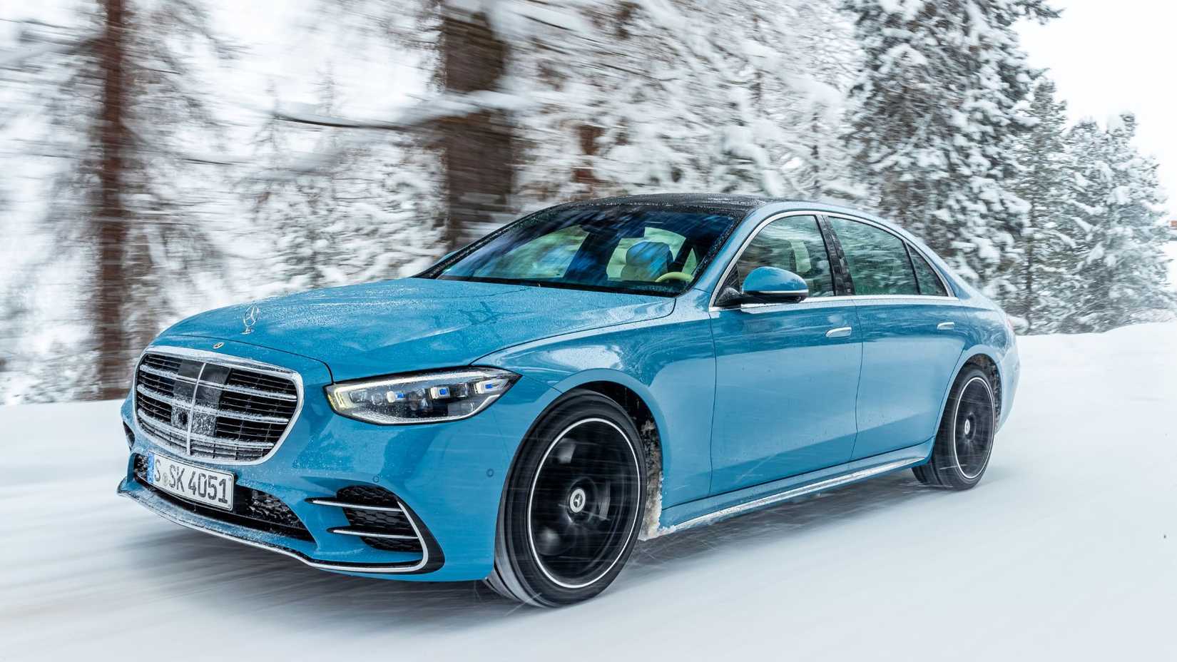 Dynamic front 3/4 shot of a blue 2024 Mercedes-Benz S-Class driving on a snowy road.