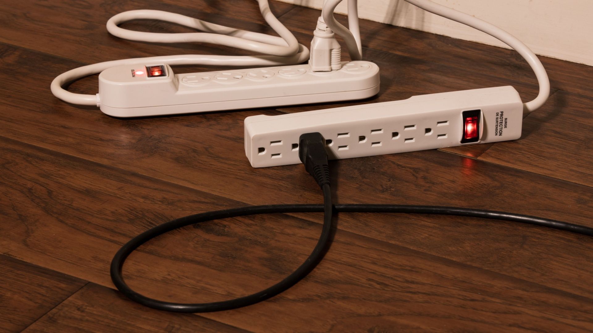 A black power cord plugged into a surge protector which is plugged into another. Known as daisy stripping, this usage of power strips is a fire hazard and is highly dangerous extension cord.