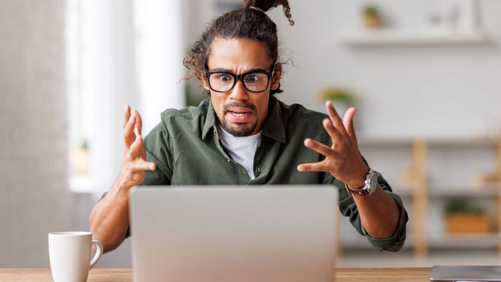 A man throws his hands in the air in frustration at his laptop.