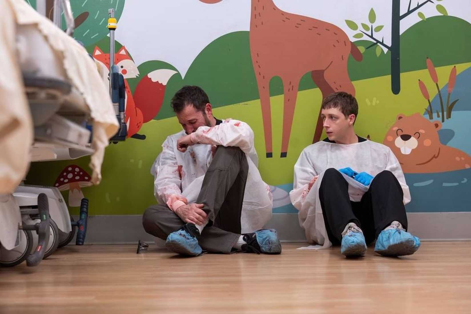Noah Wyle y Gerran Howell sentados en el suelo con una bata ensangrentada en una habitación de un hospital pediátrico.