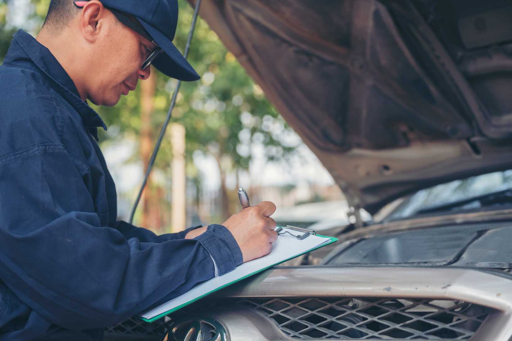 Mechanic looking under the hood with a clipboard and making notes.