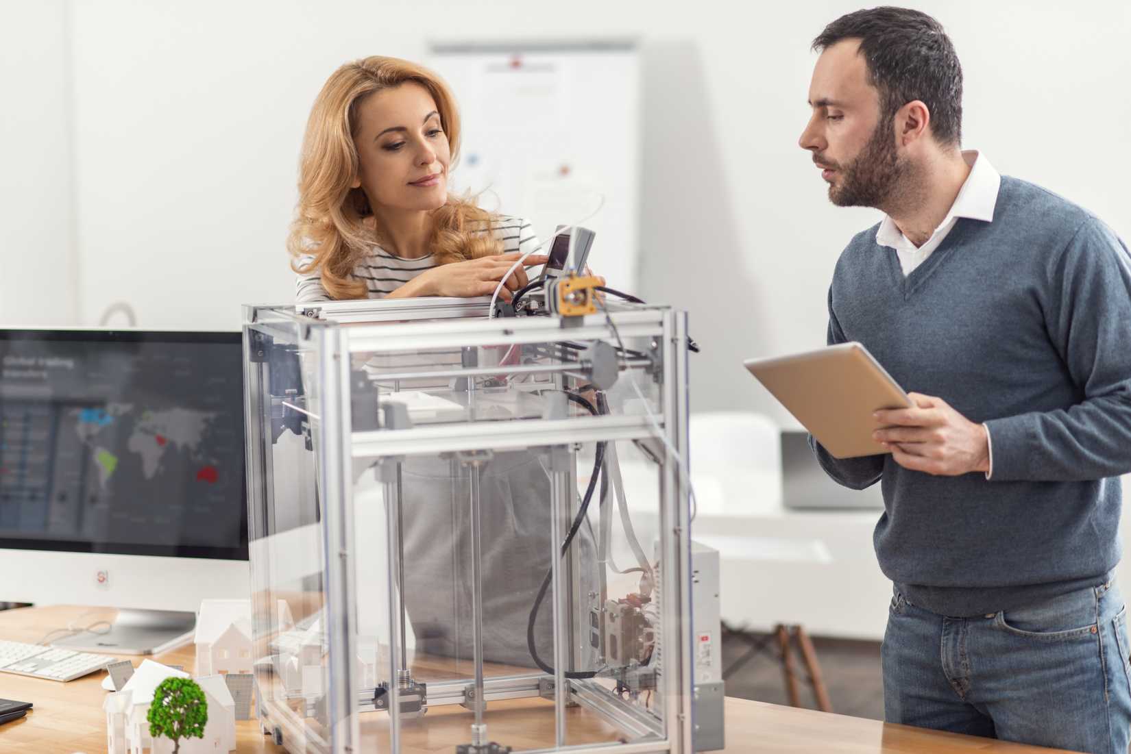 Pleasant young colleagues updating the firmware of a 3D printer while the man is controlling the process via his tablet.