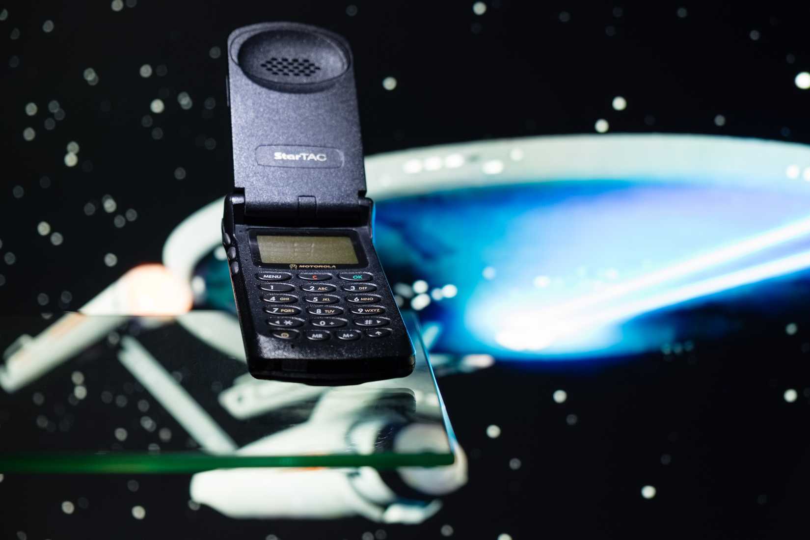 Historic GSM Motorola Startac mobile phone photographed on a glass plane with a spaceship in the background.