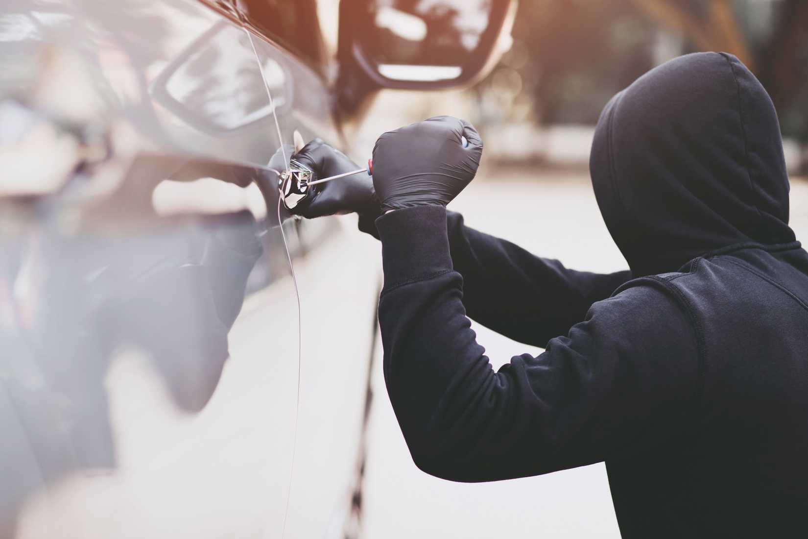 Thief in a black hoodie trying to break into a car with a screwdriver