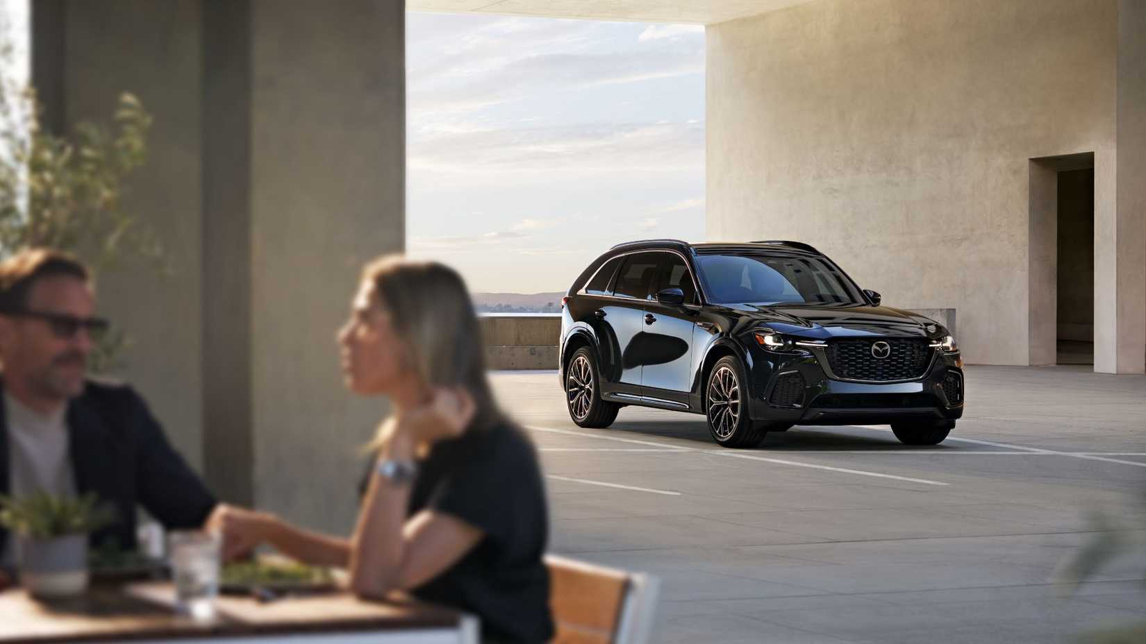 Static front 3/4 shot of a black 2026 Mazda CX-70 with people sat in the foreground.