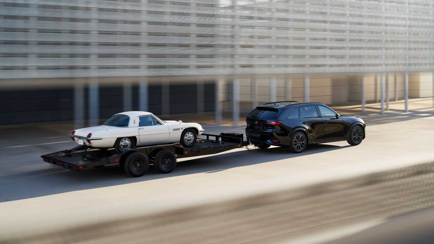 Dynamic rear 3/4 shot of a black 2026 Mazda CX-70 towing a trailer with a white car.