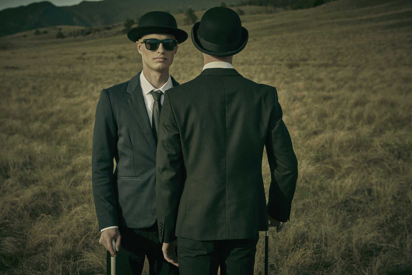 Two serious and calm men in black suits, bowler hats and black sunglasses stand facing each other in a wild open field.