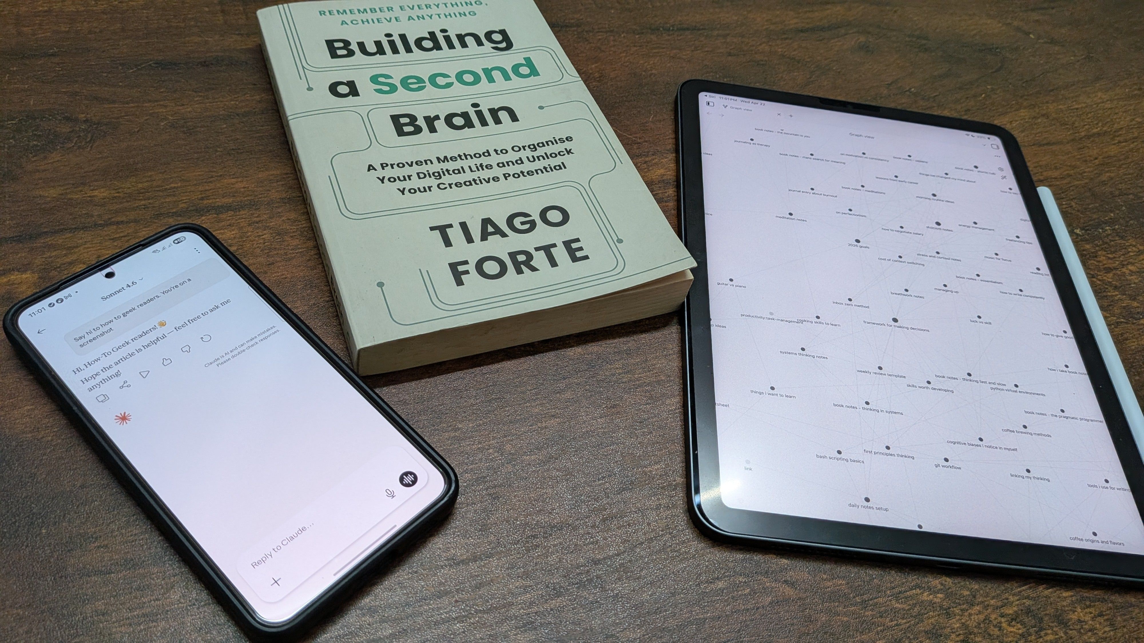A phone running Claude, an iPad running Obsidian, and Tiago Forte's Building a Second Brain book on a wooden table.