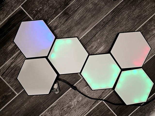 Hexagonal modular LED light panels connected together on a wooden floor, glowing in different colors with visible power and data cables.