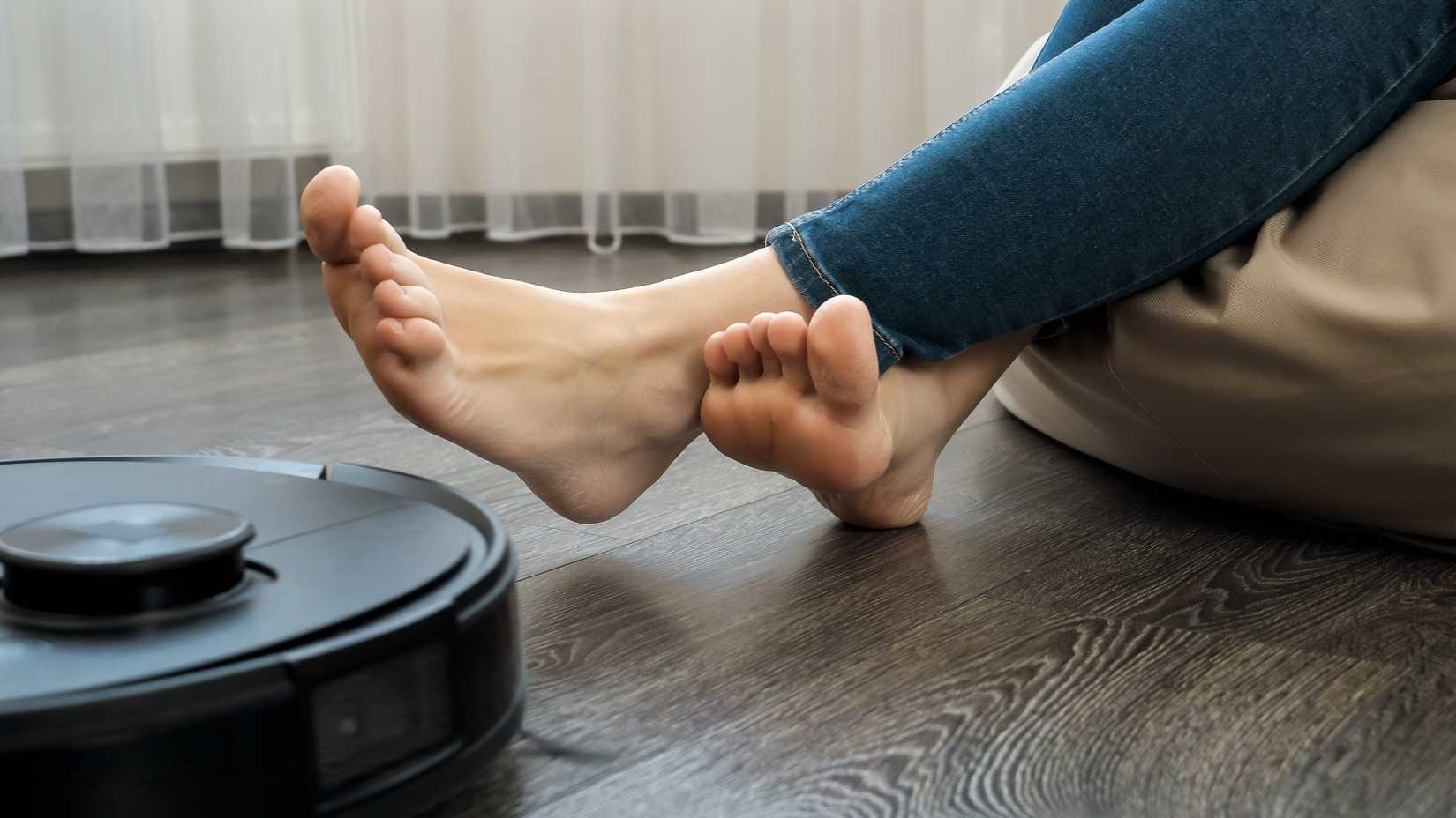 Person relaxing with bare feet on a beanbag chair while a robot vacuum cleaner moves across a hardwood floor nearby.